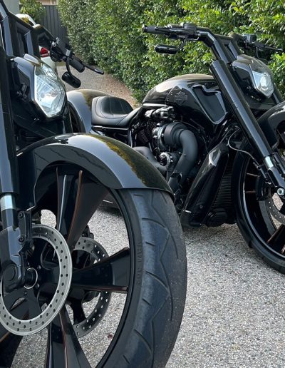 Two black custom motorcycles with large front wheels on a gravel driveway